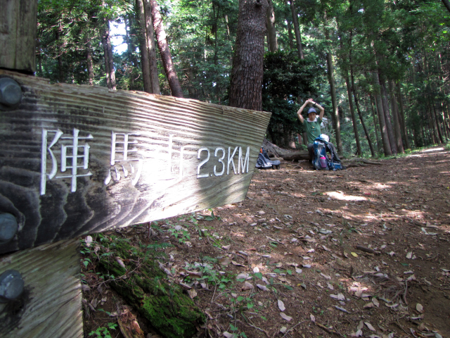 植林に囲まれた祠の手前に立つ、「陣馬山 2.3km」の標識と、休憩する隊員